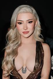 closeup portrait of a stunning blonde woman with long, wavy hair. she is wearing a lowcut, dark brown dress that reveals some cleavage.  she has professional makeup and a small tattoo visible on her chest. the background is dark and out of focus, drawing attention to the woman. her expression is serious and confident. the overall style is glamorous and elegant.