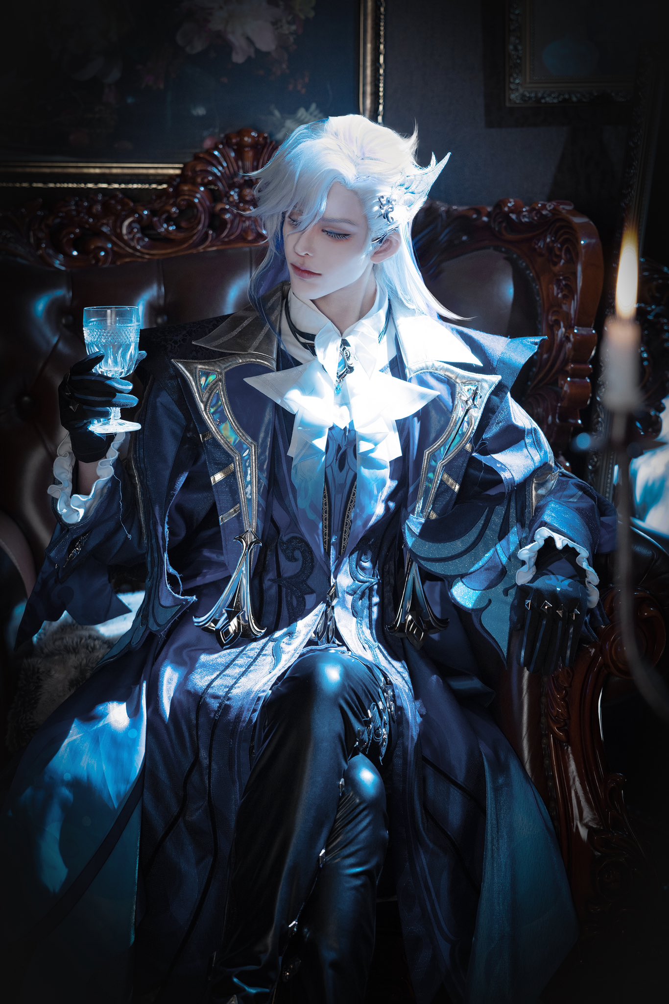 a young man cosplaying an anime character sits in a dark, ornate chair. he is wearing a blue and black costume with silver accents, and he has long, silver hair. he is holding a glass of water in his left hand.