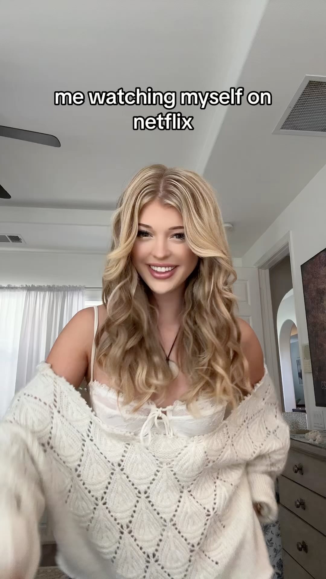 a young woman, loren gray, is smiling towards the camera while wearing a white top and a white, crochetedstyle sweater. the caption above her head says, "me watching myself on netflix." the background appears to be inside a house, likely a bedroom, with a curtain, dresser, doorway, ceiling fan, and light fixture visible.