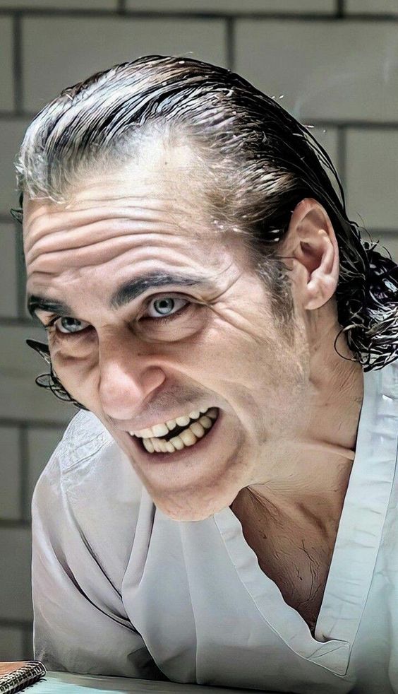 a closeup shot of joaquin phoenix as the joker in the film "joker". he is wearing a white asylum gown and leaning forward, with a manic grin on his face and a crazed look in his eyes. the background consists of white tiled walls, suggesting the setting of a mental institution.