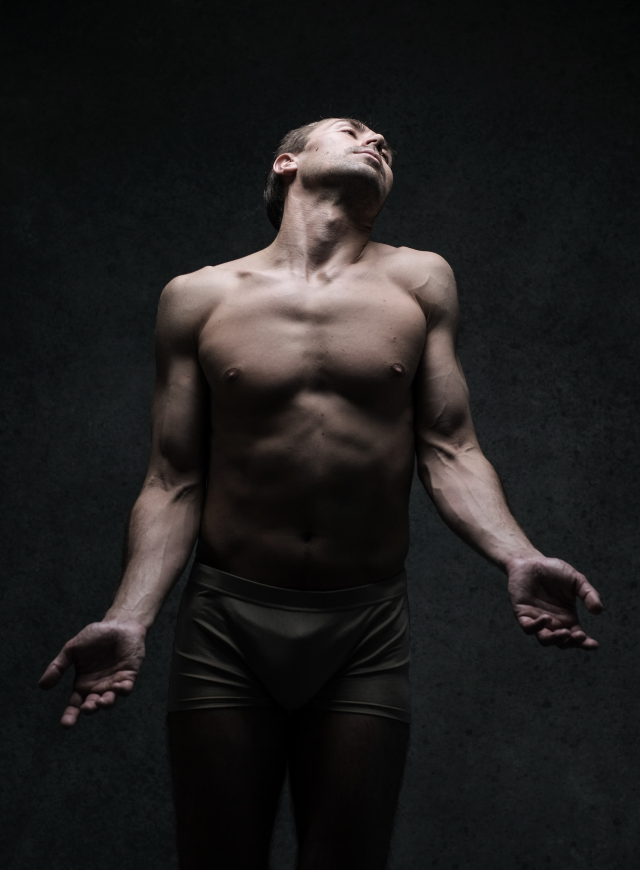 a nude man is standing in a studio setting, his arms outstretched with palms open and head tilted back. he is lit by a spotlight from above, highlighting his muscles and creating dramatic shadows on his body. the background is a dark gray.