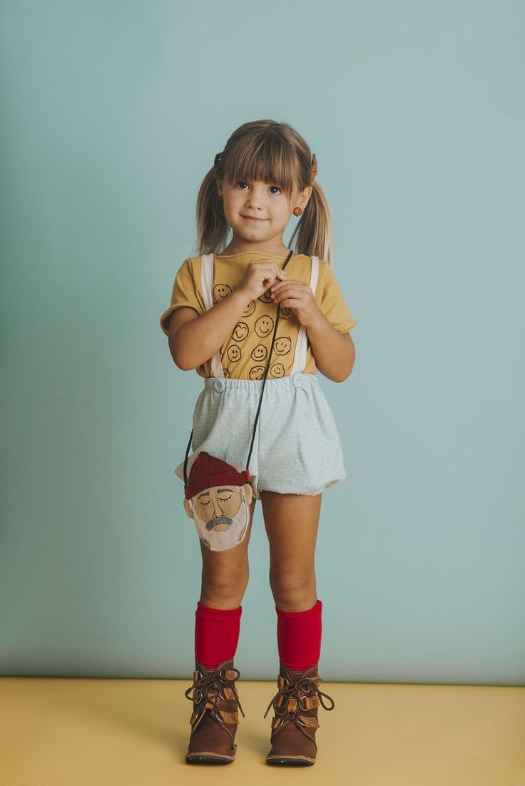 a fullbody studio shot of a cute little girl with pigtails. she is wearing a mustard yellow tshirt with smiling faces printed on it, light blue shorts, red kneehigh socks, and brown laceup boots. she's carrying a unique small handbag featuring a whimsical face. the background is a soft pastel blue, and the overall style is playful and adorable. the image highlights children's fashion with a focus on charming details.