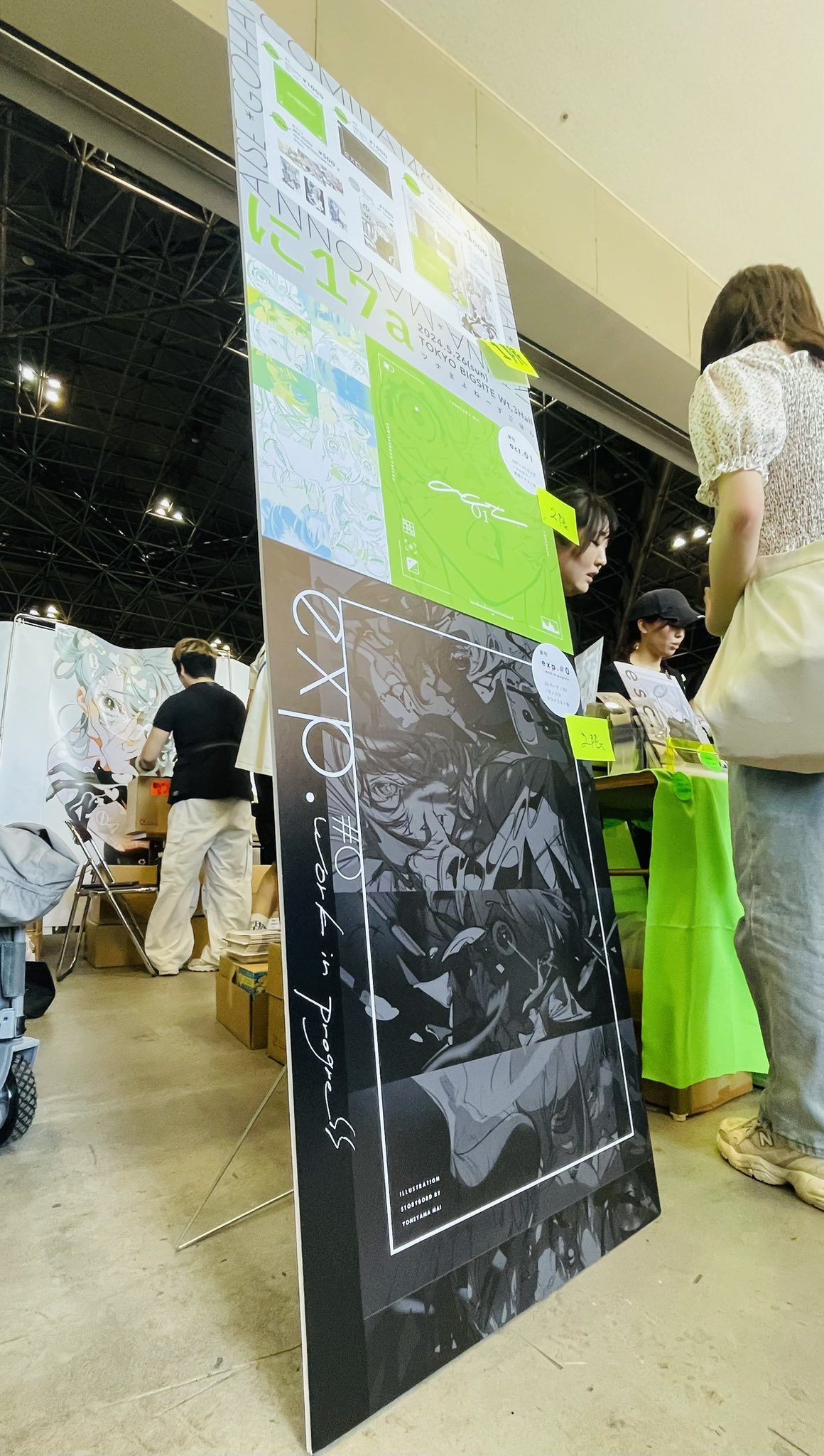 a large poster stands on the floor showcasing animestyle artwork in black and white with green accents. the top portion features colorful character designs. people are browsing at the booth, some wearing face masks.  the setting appears to be a convention hall with bright lighting and a concrete floor.