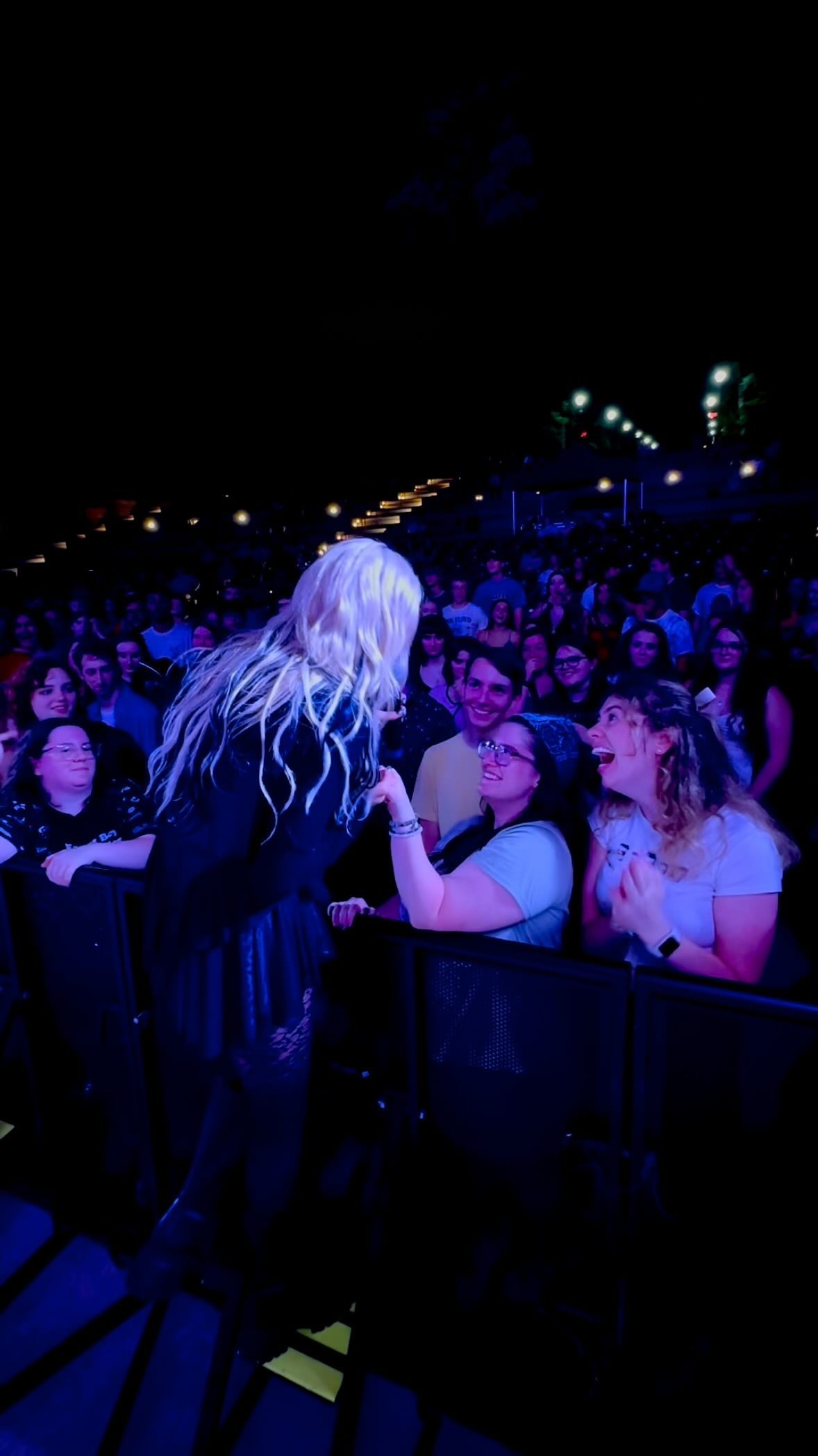a performer with blonde hair is interacting with fans at a concert. the crowd is visible in the background, and the scene is lit with blue stage lighting. the fans look excited.