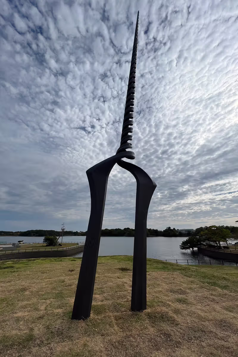 a tall, black, abstract metal sculpture stands in a park. it is a tall, thin, spiky sculpture that appears to be made up of two long pieces of metal. the sculpture is silhouetted against a cloudy sky. there is water, likely a lake or a pond in the background. the ground is covered in grass.