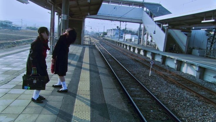 the image shows two japanese schoolgirls in winter uniforms standing on a nearly deserted train platform in japan.  one girl is looking down, while the other is leaning back with her head tilted up. they both appear to be carrying school bags.  the platform is long and stretches into the distance, showing railway tracks leading towards a grey, overcast sky. the overall atmosphere is quiet, lonely, and somewhat melancholic, suggestive of a cold winter day and perhaps a feeling of isolation or waiting.  the scene is set at a seemingly rural train station, with buildings and fields visible in the background.