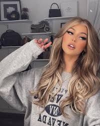 a woman with blonde, wavy hair is wearing a grey sweatshirt and giving a peace sign. she has makeup on and is in front of a shelving unit with various accessories, including a handbag, picture frame, and backpack.