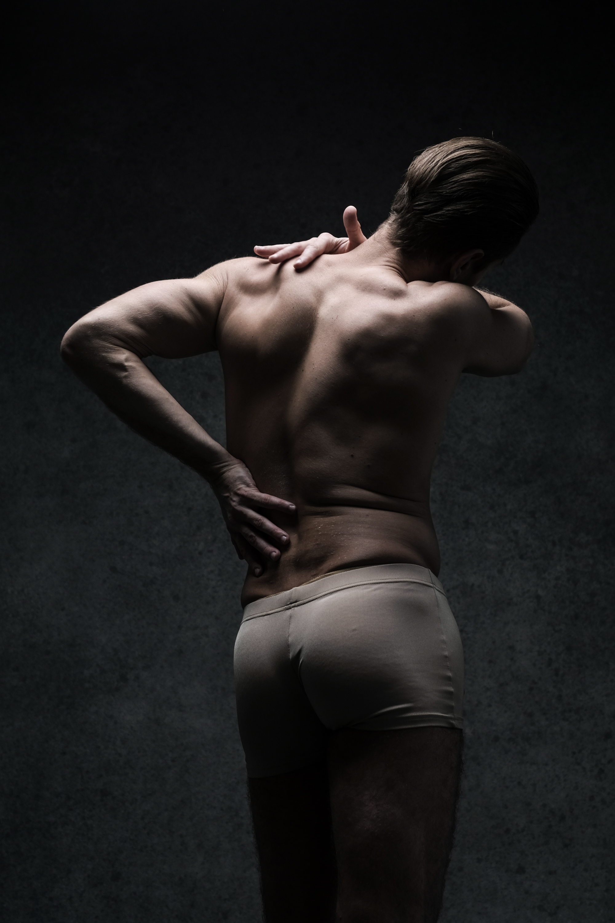 a shirtless man stands with his back to the camera, clutching his lower back in apparent pain. his posture suggests discomfort and he is illuminated by dramatic studio lighting against a dark background.