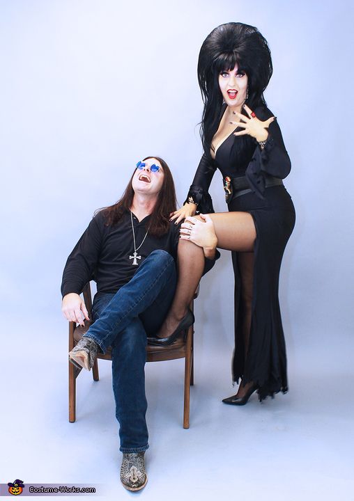 a woman dressed up in a long black dress with a tall black wig, smiles at the camera as she poses with her hand on the shoulder of a man dressed in a black shirt and blue jeans, who is laughing while sitting in a wooden chair. the two are dressed in a spooky/halloweenthemed style and pose in front of a plain white background.