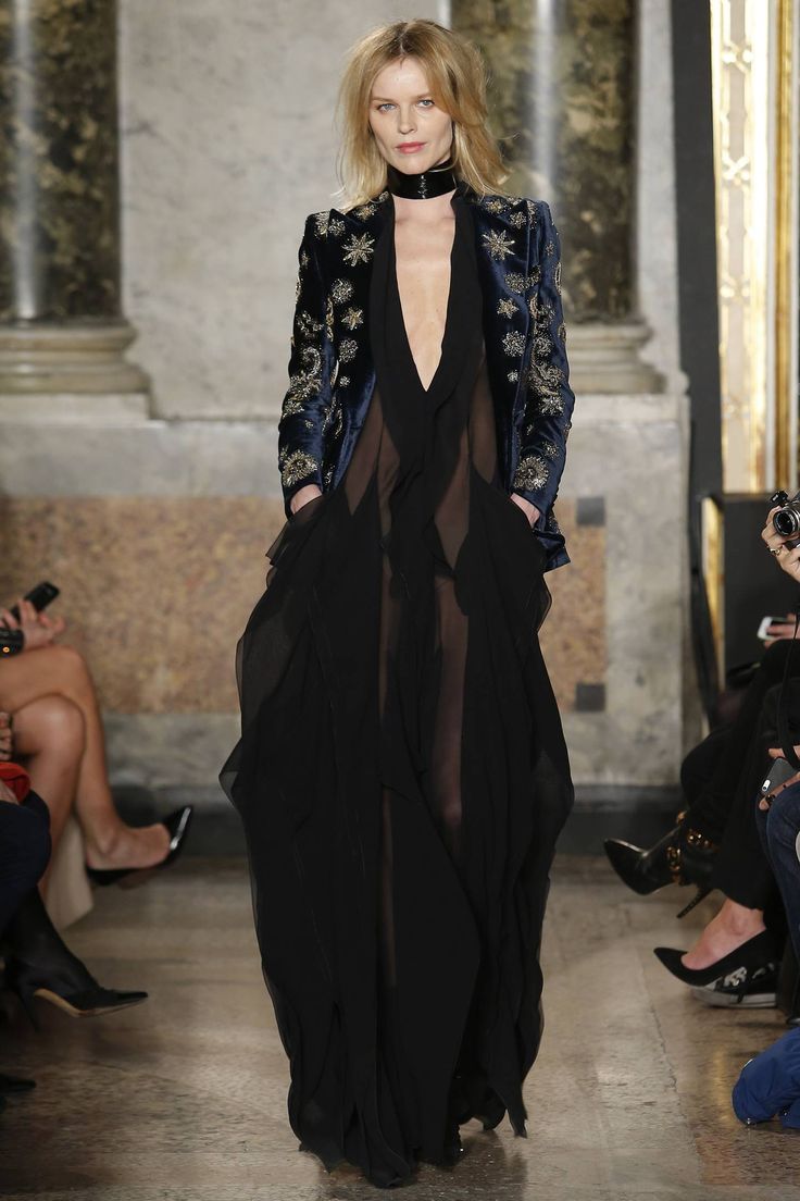 a fulllength shot of a fashion model walking down a runway during a fashion show. the model is wearing a black sheer maxi dress layered over a dark navy velvet jacket adorned with starshaped embroidery. she accessorizes with a black choker necklace. the background features a marble interior with pillars, providing an elegant backdrop to the fashion show.  other people's feet and legs are visible in the background, suggesting a crowd observing the runway show. the model's pose and clothing exude elegance and sophistication, typical of highfashion photography.