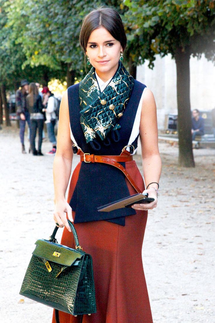 a stylish brunette woman is walking down a treelined parisian street. she's wearing a sophisticated outfit consisting of a dark blue vest over a rustcolored midi skirt, a green silk scarf, and a wide brown belt.  she's carrying a dark green hermès kelly bag made of crocodile skin.  her accessories include emerald earrings and a simple necklace. the overall style is elegant and parisian chic. the image captures a moment of street style fashion.