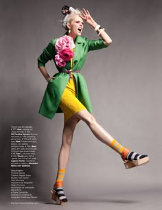 a woman in a green jacket and a yellow skirt poses in the studio. she has a flower accessory in her hair and is wearing colorful socks and shoes.  the background is a simple gray.