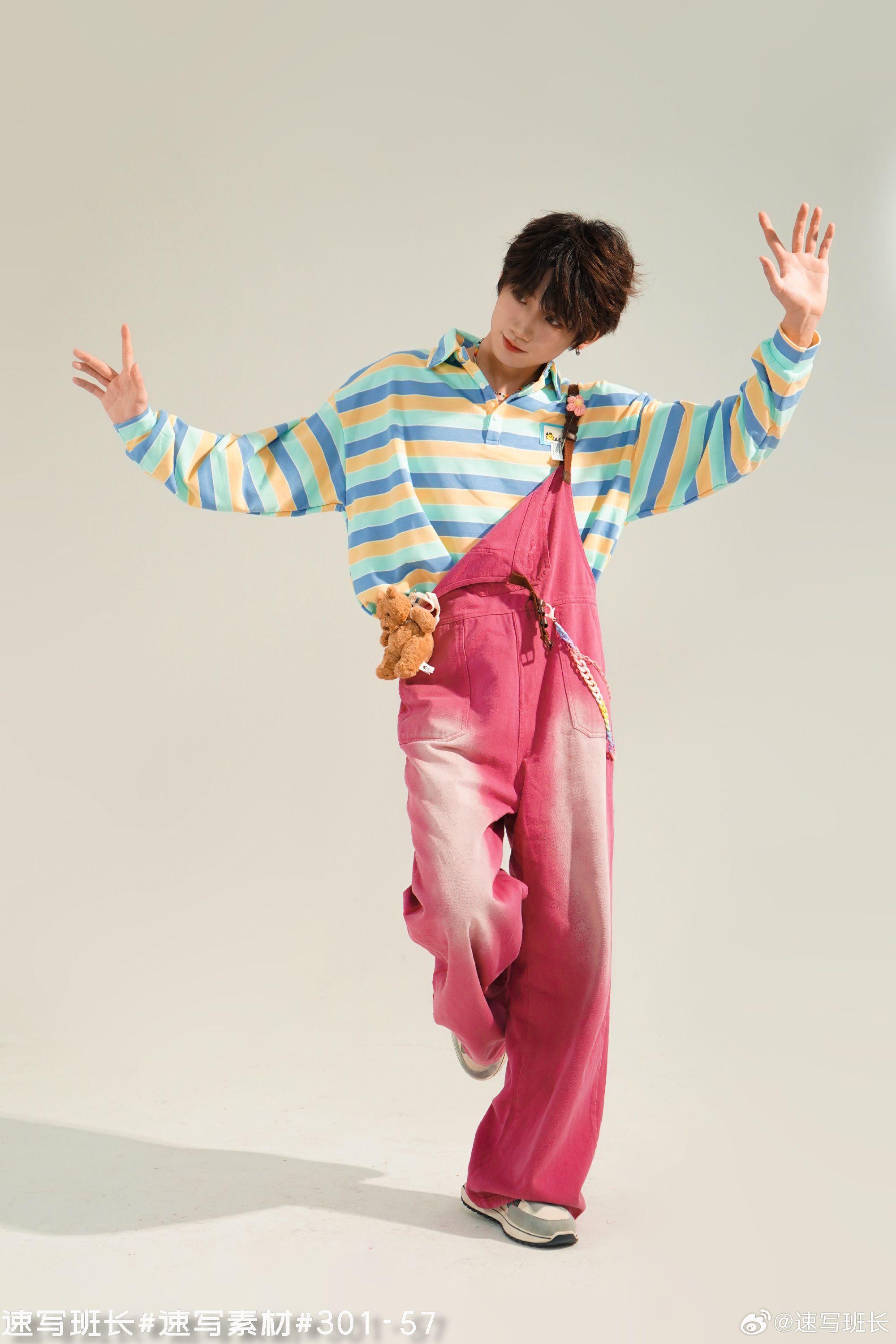 a young asian man strikes a playful pose in a studio setting. he's dressed in a colorful outfit featuring a striped, pastelcolored shirt and bright pink overalls. a small teddy bear peeks out from the front pocket of the overalls. the lighting is bright and even, characteristic of a professional studio environment.