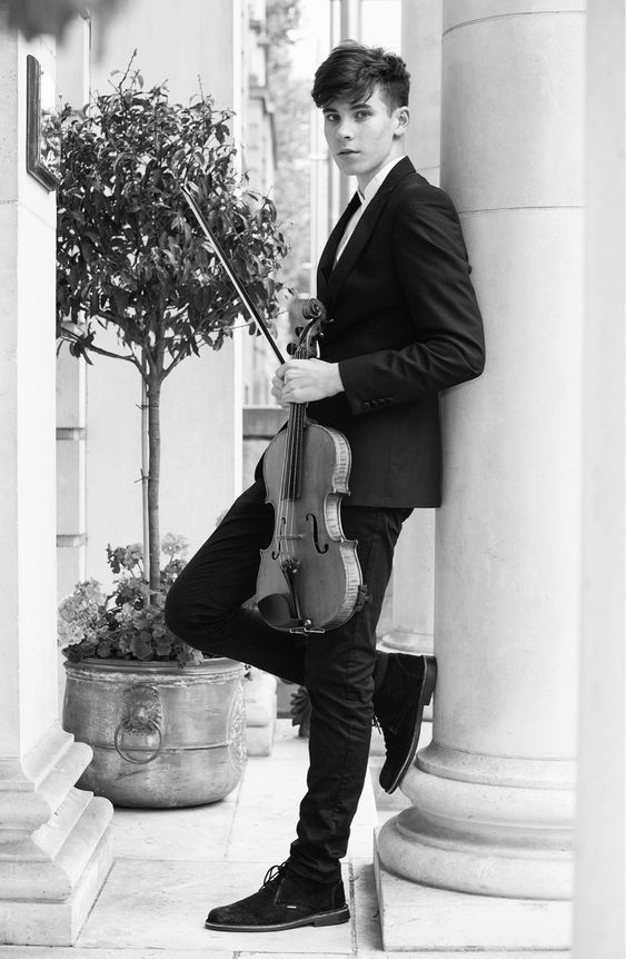 a black and white photo of a young man dressed in a suit, standing outdoors and leaning against a pillar. he's holding a violin and looking directly at the camera with a serious expression. the background suggests a sophisticated urban environment with classical architecture. there's a potted plant to the left. the lighting is soft and natural, casting subtle shadows.