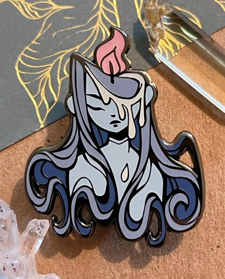 a closeup shot of an enamel pin depicting a woman's head and shoulders, stylized as a candle. the woman has a gray complexion, a light gray face, and darklined closed eyes and a downturned mouth. dripping white wax covers her forehead and flows down her face. on top of her head is a small, stylized pink flame. the pin has a dark gray, curled hairlike design around her face and shoulders, resembling dripping wax. in the background, there are crystals and a gold leaf pattern on a gray surface. the pin is placed on a brown surface.