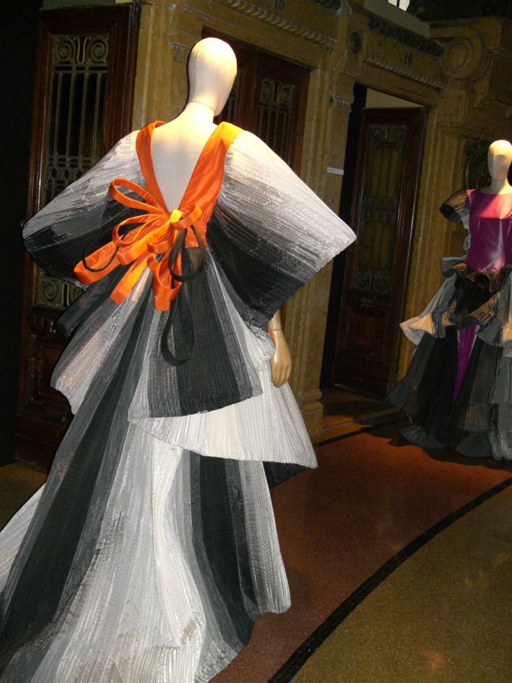 the image shows a mannequin wearing a dress with a black and white gradient and an orange ribbon on the back. the mannequin is standing in an exhibition hall with other mannequins and dresses visible in the background.