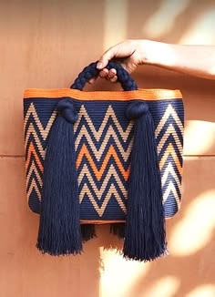 a close up shot of a woman's hand holding a blue, white and orange knitted boho bag with tassels. the bag is colorful and patterned with a zig zag design. the image is taken against a brown background.