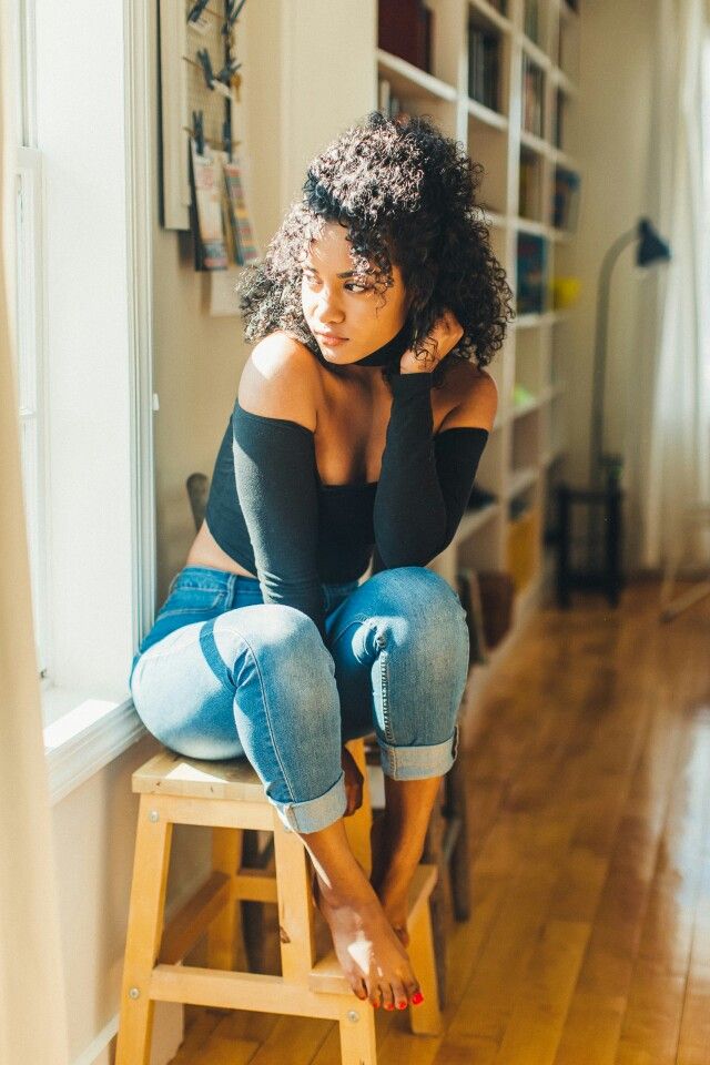 a young woman with curly hair sits on a wooden stool in front of a sunlit window. she is wearing a black offtheshoulder top and blue jeans. she has a thoughtful expression on her face and is looking away from the camera. the room she is in is minimally decorated with a bookshelf in the background.