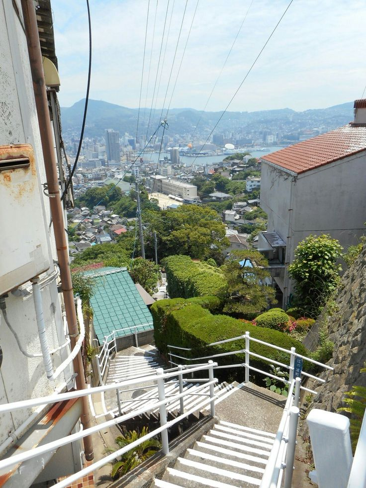 a highangle shot looking down a long staircase with white railings, offering a scenic view of a cityscape with buildings, trees, and a harbor in the background. the surrounding area consists of houses and greenery, adding to the urbanmeetsnatural landscape. overhead, power lines crisscross the sky, and a distant hill or mountain is visible in the horizon.