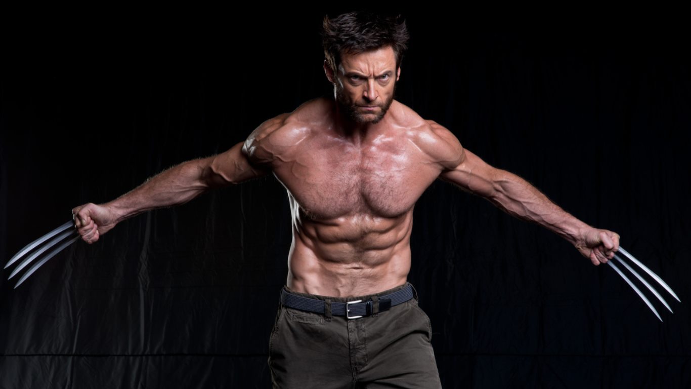 the image shows hugh jackman posing as wolverine, a character from marvel comics. he is shirtless, displaying his muscular physique and defined abs. he is holding wolverine's signature metal claws in both hands, and has a serious, determined expression on his face. he's wearing dark pants with a belt. the background is black, suggesting a studio shot.