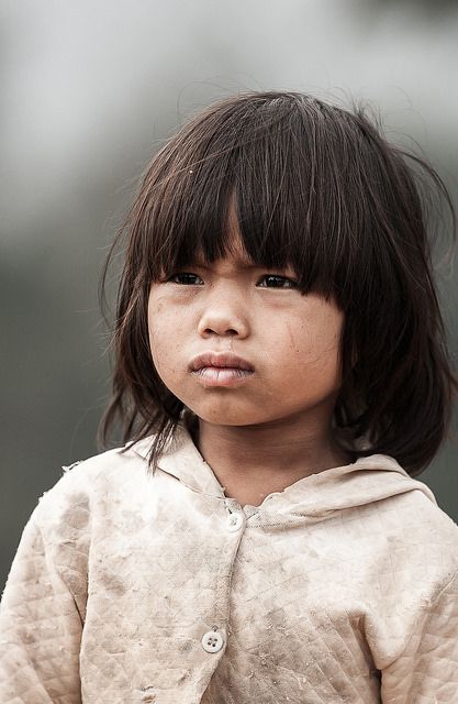 a young asian girl with a serious expression is captured in a portrait. she has dark hair with bangs and is wearing a dirty, worn jacket with buttons. her face appears slightly dirty, and she is looking away with a sad, innocent gaze. the overall impression is one of poverty and hardship.