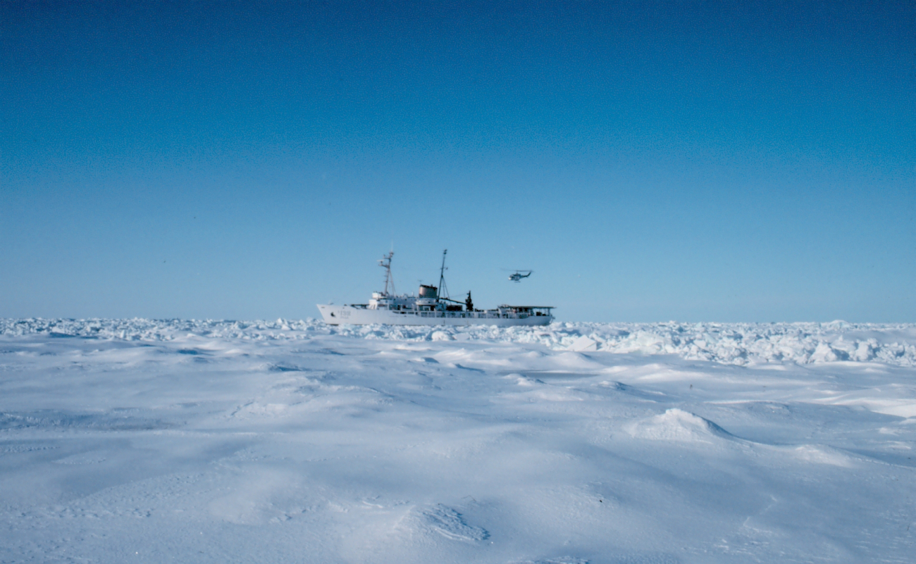 a white ship sails on a frozen sea under a clear blue sky. there is a helicopter flying above the ship. the ice floes are very large and cover the entire surface of the sea.