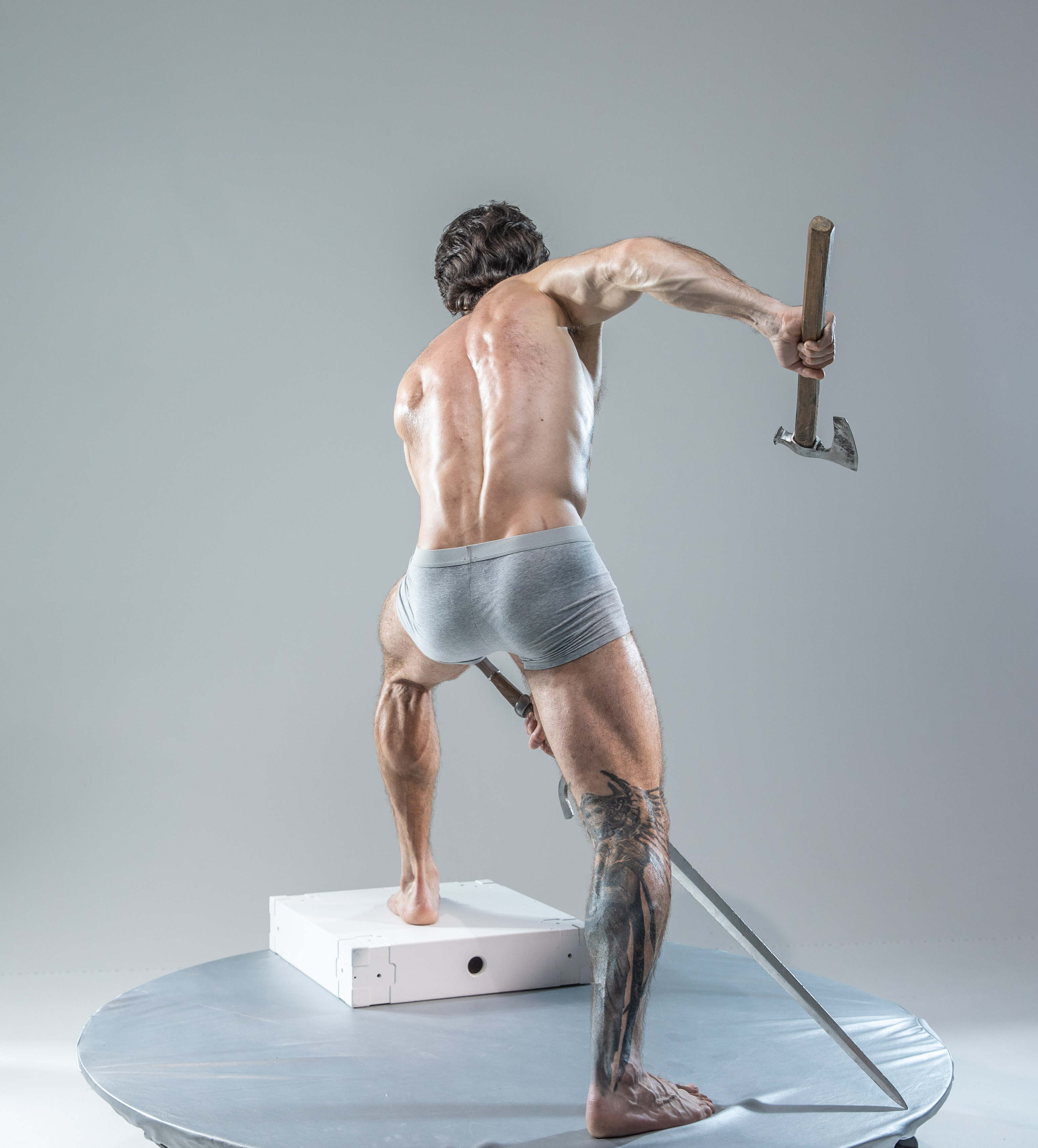 a muscular man with his back turned to the camera, holding an axe in his right hand and a sword in his left. he is in a fighting pose, standing on a white platform with a gray background.