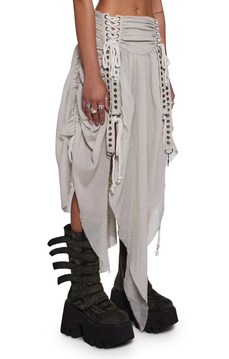 a fullbody studio shot showcases a model wearing a unique, asymmetrical beige skirt.  the skirt is multilayered, with draping fabric and features a drawstring waist, corsetlike lacing detail at the top, and metal studs and rope accents down the sides. the model also wears dark green chunky platform boots with multiple buckles.  the overall style is a blend of grunge, boho, and alternative fashion.  the model's hands show several rings.