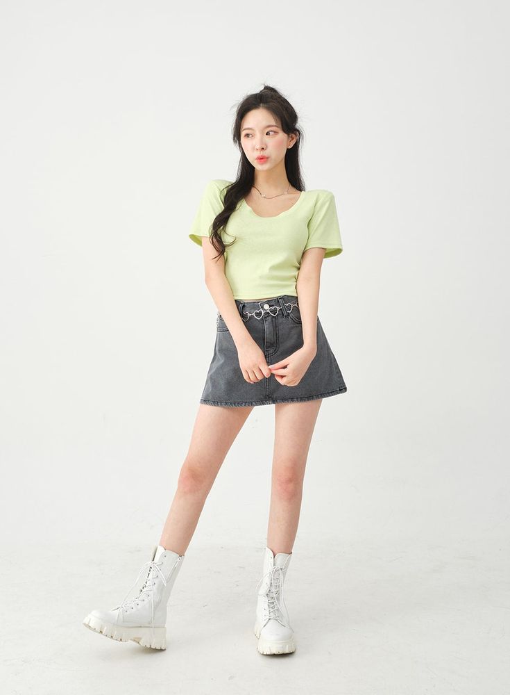 a young asian woman is standing against a white background, showcasing a stylish outfit. she is wearing a light green shortsleeved top and a dark grey denim mini skirt with a heartshaped chain belt. the skirt hits her midthigh, and she’s wearing white laceup combat boots. her hair is pulled back into a high ponytail, and she appears to be posing for a fashion shoot. the overall style is trendy and casual, perfect for everyday wear.