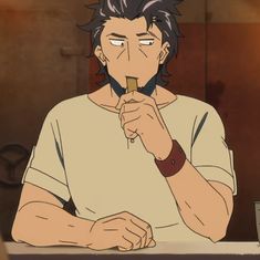 a man with short, dark hair and stubble is pictured from the chest up. he is indoors and is wearing a loosefitting, shortsleeved, light brown shirt and a dark brown wristband on his left wrist. he is eating a small, rectangular snack, holding it between his thumb and index finger. the background is out of focus, but appears to be a room with light brown walls. the overall color palette is warm and muted.
