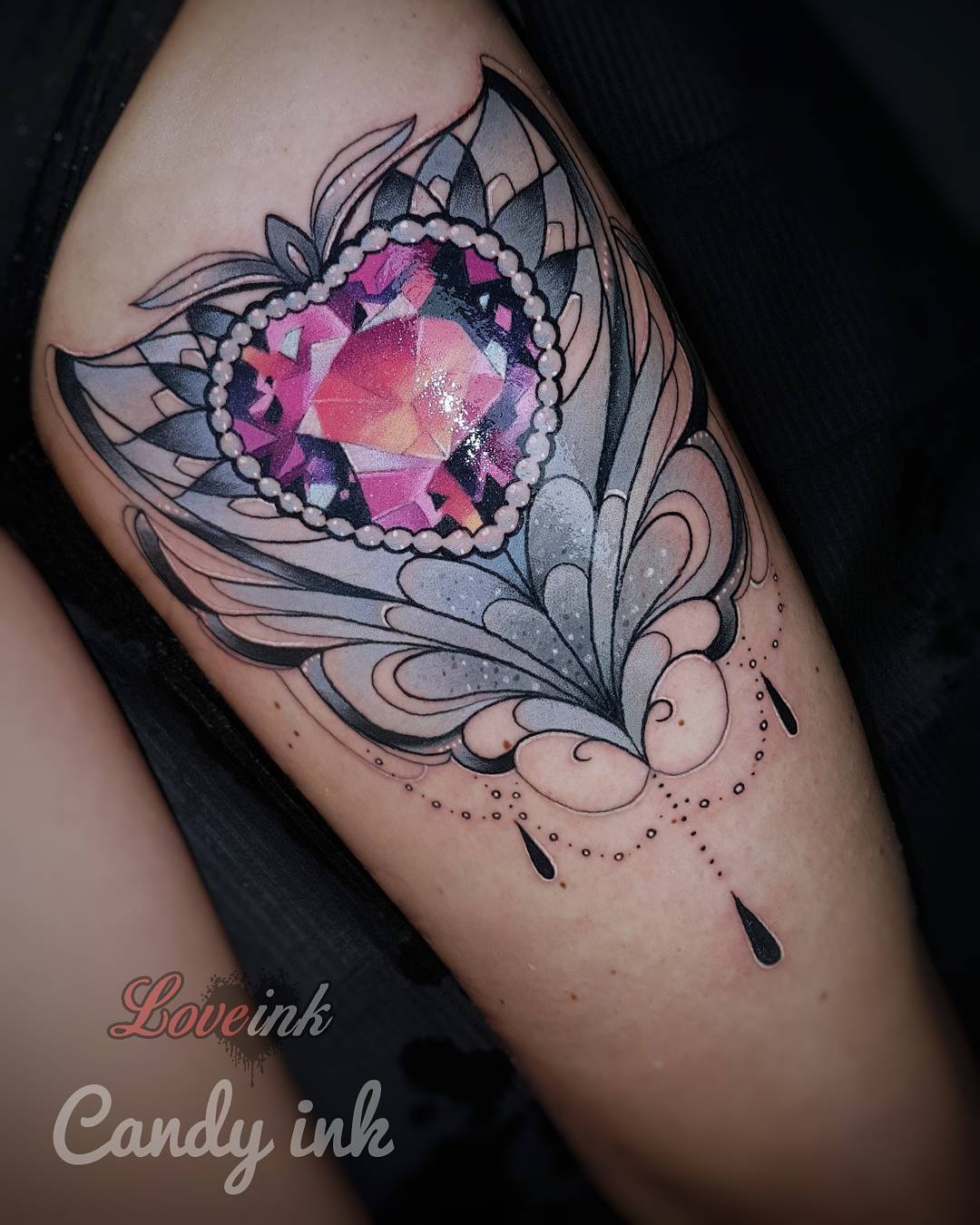 a closeup photograph showcasing a freshly done thigh tattoo. the tattoo features an intricate, stylized heartshaped jewel rendered in shades of pink and purple. it is encased in a decorative, blackwork design reminiscent of lace or filigree, which extends outwards and downwards, ending in delicate droplets. the tattoo sits on the outer thigh of a person, whose skin appears lightly tanned. the surrounding environment suggests a tattoo parlor setting with soft, artificial lighting highlighting the tattoo's details.