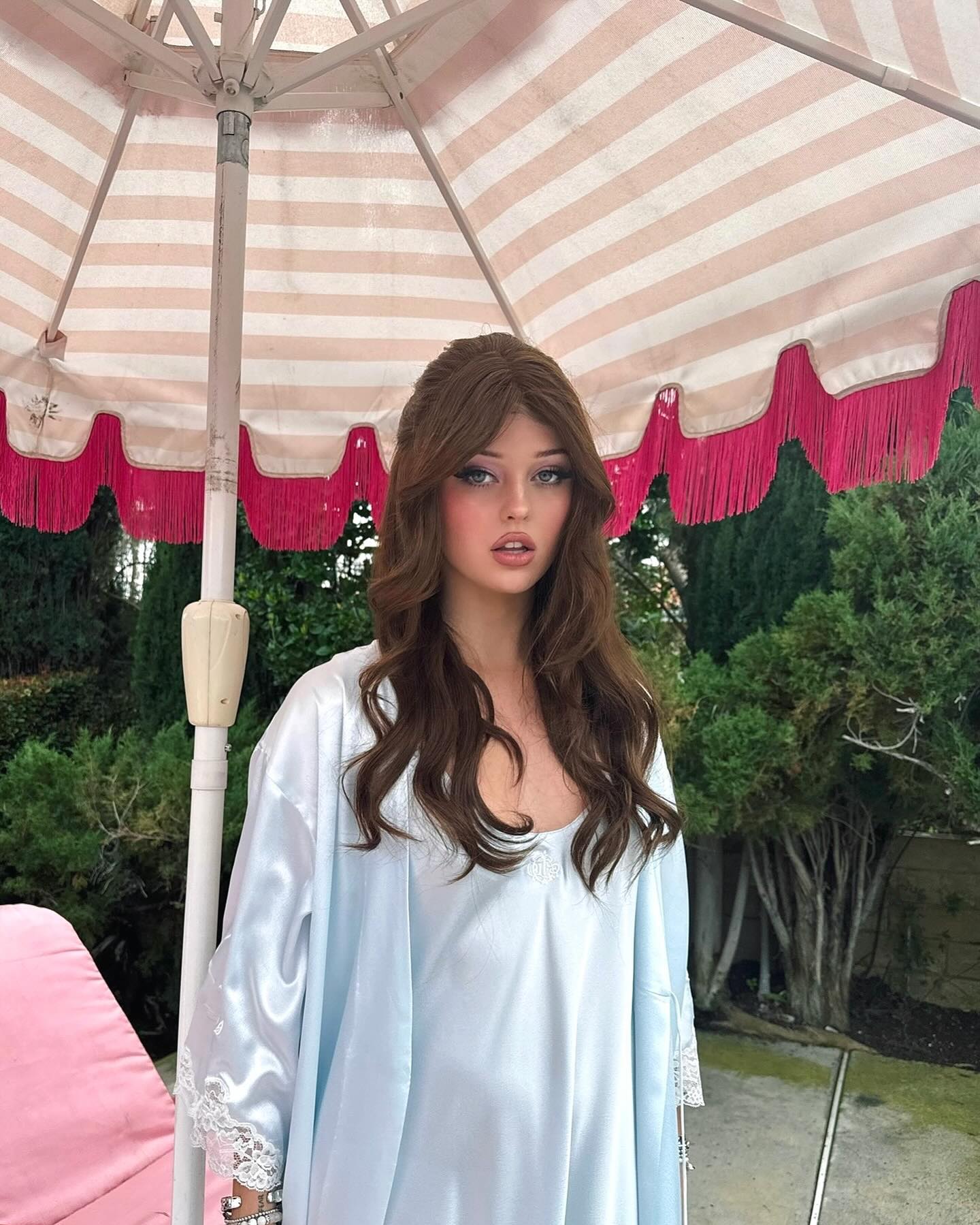 a woman with long brown hair is standing under a pink and white parasol with green bushes and trees in the background. she is wearing a light blue satin robe with lace trim. she is wearing heavy eye makeup.