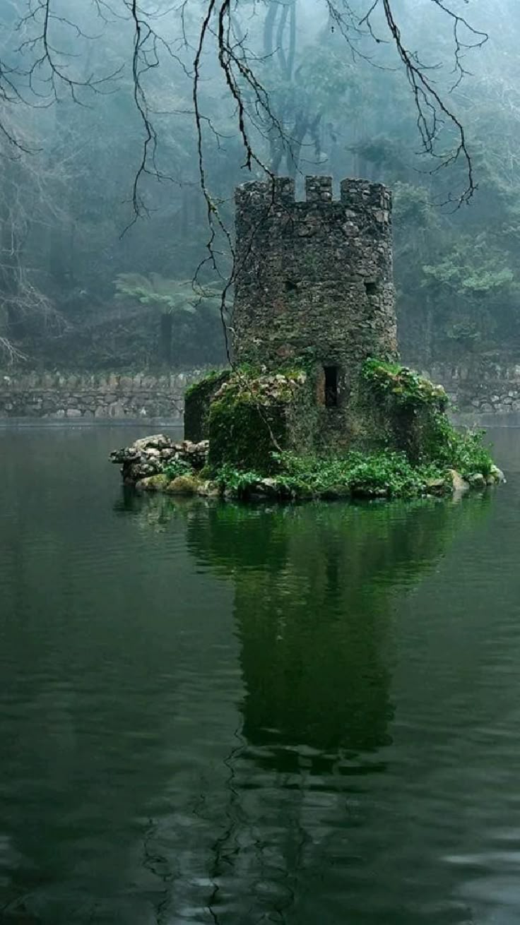 the image shows a small stone tower situated on a mossy island in the middle of a lake. the tower is reflected in the still water. the background is a misty landscape with trees. the scene evokes a sense of serenity and historical charm.