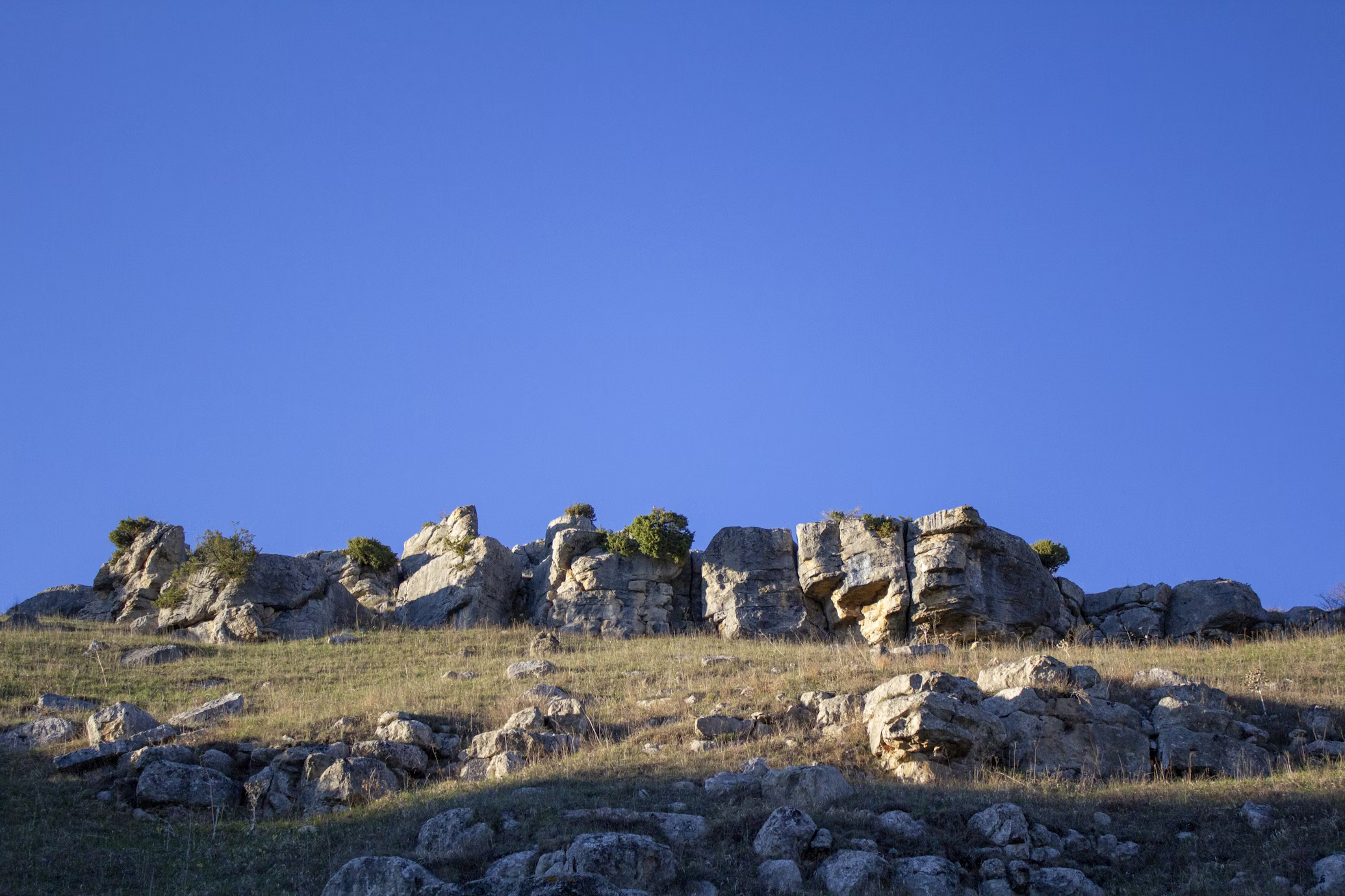 a blurry photo of a hill with rock formation on top against a bright blue sky. the hill is covered with dry, yellow grass and some green bushes. the photo appears to be taken during a sunny day, as the shadows are visible on the grass.