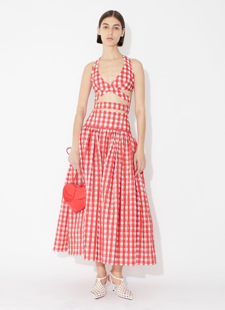 a woman stands in a studio shot, modeling a red and white gingham crop top and matching maxi skirt. she holds a red heartshaped bag in her left hand and wears white woven heels.