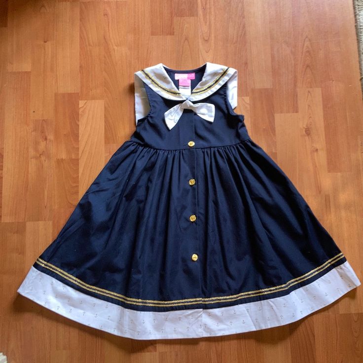 a navy blue and white sailor dress for girls is laid out on a wooden floor. the dress has a white sailor collar and a white bow at the neck. the dress has a gold trim and gold buttons down the front. the dress is sleeveless and has a full skirt.