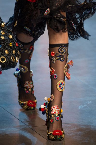 the image is a closeup shot of a model's legs and feet at a fashion show, likely for dolce & gabbana. the focus is on the elaborate footwear and hosiery. the model wears sheer black socks heavily embellished with colorful floral embroidery, beads, pompoms, and other textile details.  the shoes themselves are highheeled sandals, similarly adorned with rich, textured embroidery. the model's dress is a black lace garment visible at the top of the image. the overall style is opulent, luxurious, and indicative of high fashion. the background is blurred, keeping the focus squarely on the intricate details of the outfit.