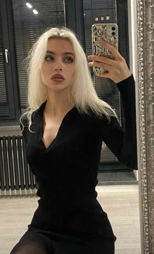 a young blonde woman is taking a mirror selfie. she is wearing a black ribbed mini dress with a vneck and long sleeves. her blonde hair is long and slightly messy. she is holding her phone in one hand and is smiling slightly. the background shows a radiator, window, blinds and a wooden floor.