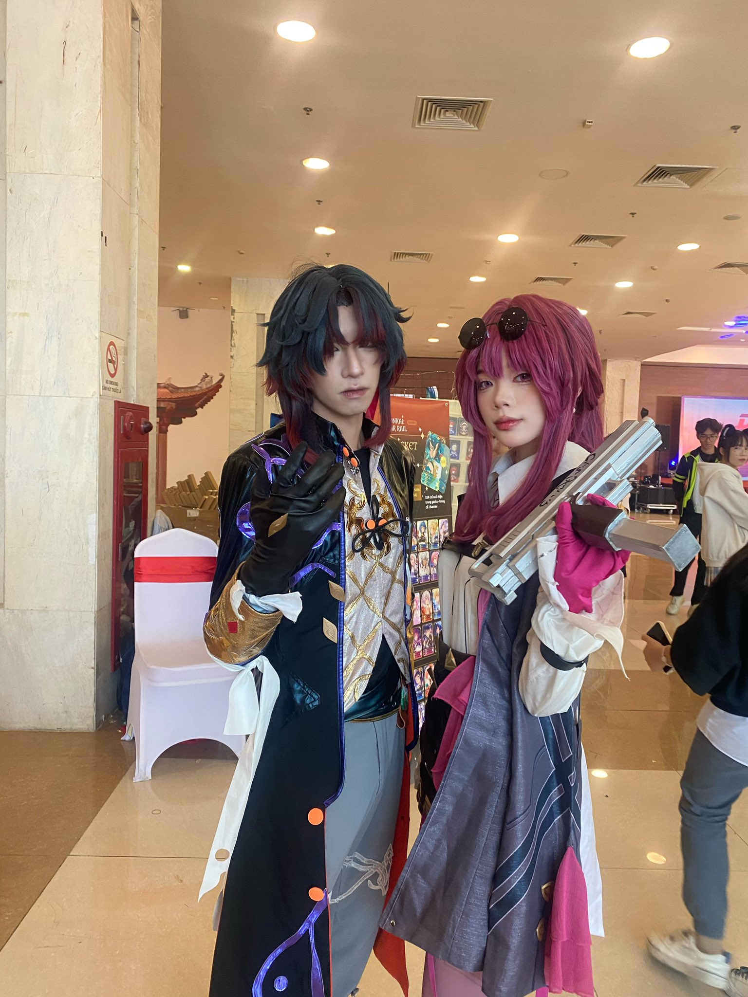 two people are cosplaying anime characters. the male cosplayer is dressed in a black and purple costume with a golden design, and he is holding a gun. the female cosplayer is wearing a purple and pink outfit with a white shirt, and she is holding a gun. they are both smiling and looking at the camera. they are standing in a room with white walls and a white tiled floor. there are some white chairs and a red and white banner in the background.