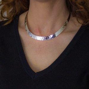 a closeup shot of a woman wearing a hammered silver choker necklace. the necklace is a statement piece, reflecting the light and highlighting the collarbone. the woman is wearing a simple black top, allowing the necklace to stand out.