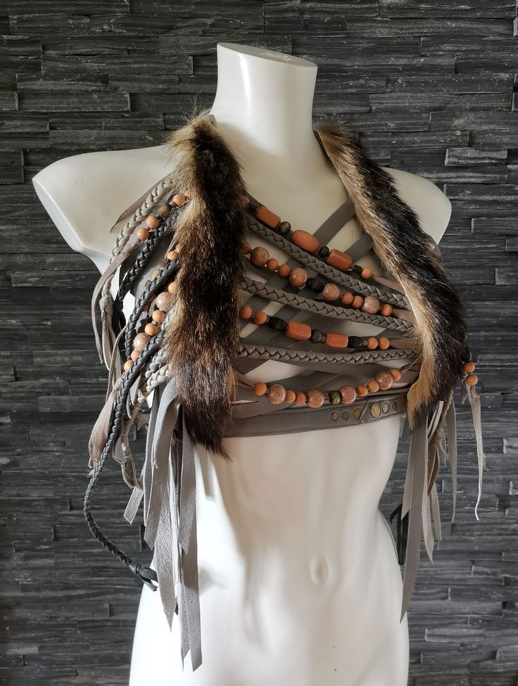 the image showcases a handmade bohostyle bralette or top.  the garment is predominantly grey and features several layers of leather or suede cords and straps.  these are interspersed with strands of beads in peach, brown and grey tones.  a significant section of dark brown fur is incorporated into the design, adding texture and visual interest.  the garment has long suede fringe that hangs below the waist. the bralette is displayed on a white mannequin against a dark grey stone background, highlighting its intricate detailing. the overall aesthetic is earthy, rustic, and evocative of tribal or festival fashion.