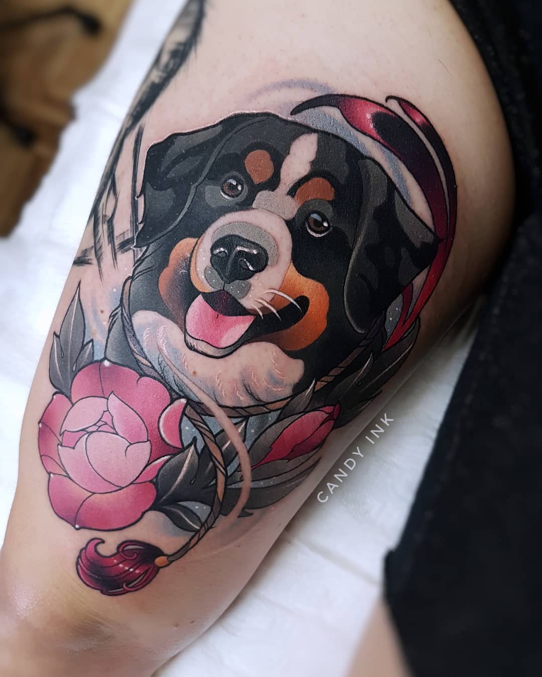 a person's thigh with a colorful tattoo of a bernese mountain dog wearing a santa hat.  there's a pink rose and rope details as well. the tattoo artist's signature "candy ink" is visible in the image.