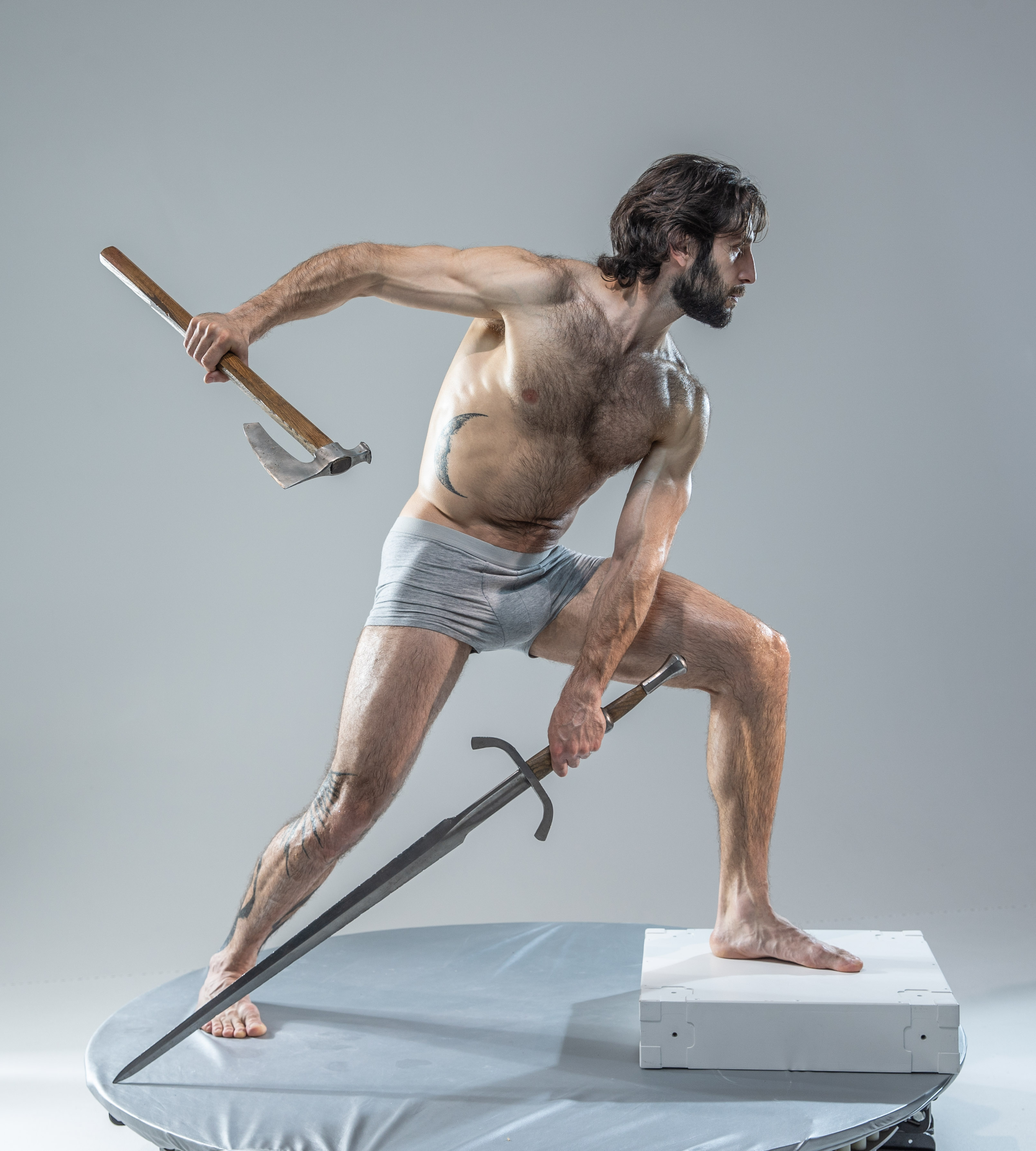 a muscular, bearded man stands barefoot on a round platform in an action pose, wielding an axe in his right hand and a sword in his left. he is wearing grey underwear and his body is angled, suggesting movement. the background is a neutral grey, illuminated with soft light. his weapons appear to be crafted from metal and wood.