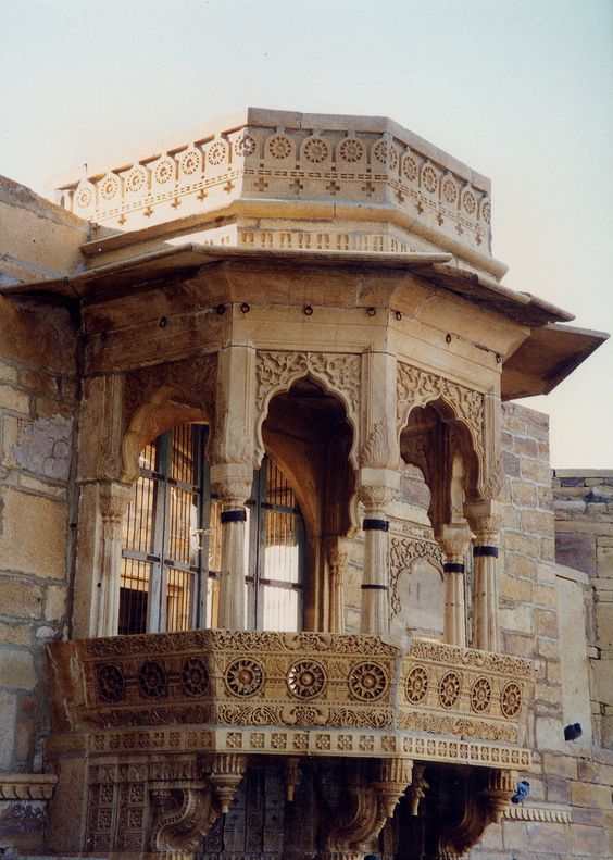 this is a detailed closeup shot of an ornately carved stone balcony, likely found in jaisalmer, india. the balcony features intricate jali work, a type of latticed screen common in indian architecture, and is made entirely from a pale sandstone. the image captures the fine craftsmanship and traditional architectural style of the region.