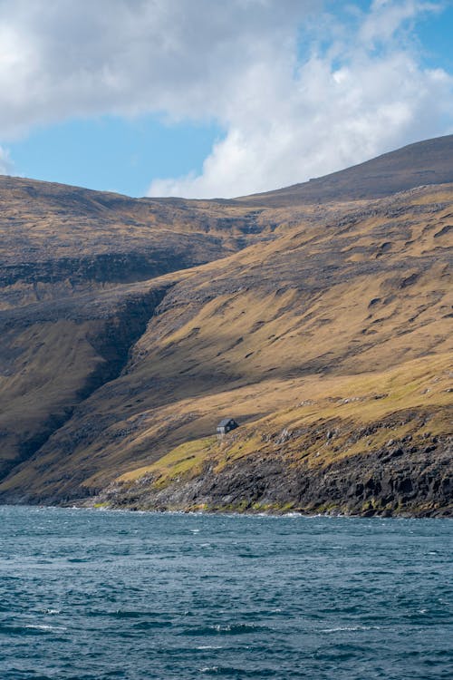 a beautiful landscape photo showing a single house nestled into the side of a large hill. the hill slopes down to a rocky shore which ends at the deep blue water of the ocean. the sky is bright blue with large white clouds.