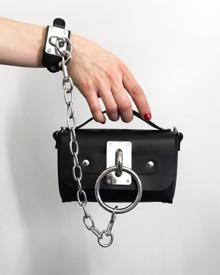 a person is holding a black leather purse with a chain and metal accents. the chain is connected to a bracelet on the person's wrist, and a large metal ring is attached to the front of the purse. the person has red nail polish on and is holding the purse by the top handle.