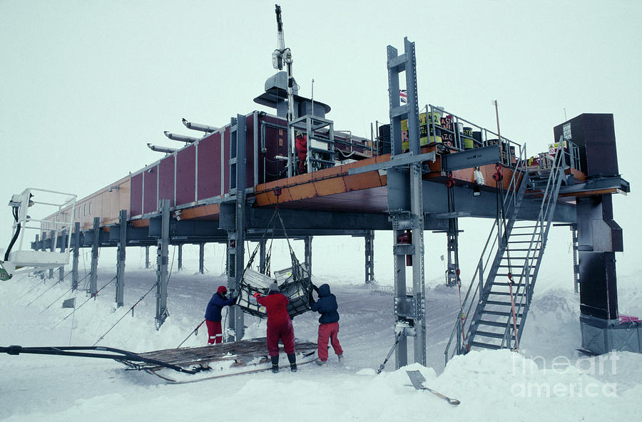 a view of a research station in antarctica, likely mcmurdo station.  the station is built on stilts,  and there are three scientists in the foreground, working to lift supplies from a wooden sled up to the station.  the station is made up of multiple modules that are connected together.  a crane is shown lifting a crate up to the station.  a set of stairs leads up to the station.  the snowy landscape suggests this was taken during the winter months.
