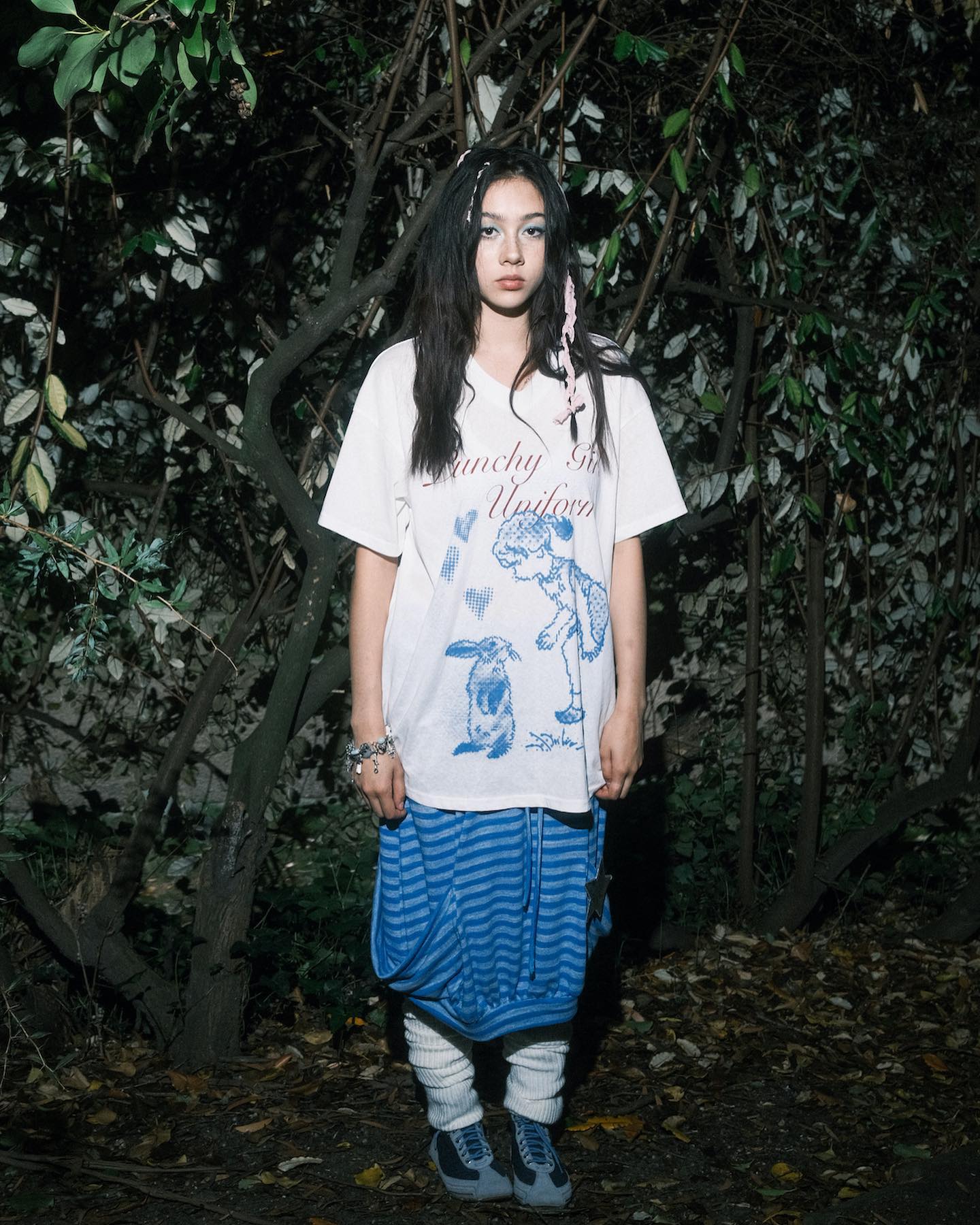 a young woman stands in a dimly lit forest setting. she is wearing a white graphic tshirt with a whimsical illustration, a blue and white striped skirt, white socks, and blue sneakers. her long dark hair is styled simply, and she has minimal makeup. the overall style is alternative and slightly grunge, with a touch of whimsicality from the tshirt's design. the image has a moody, atmospheric feel.
