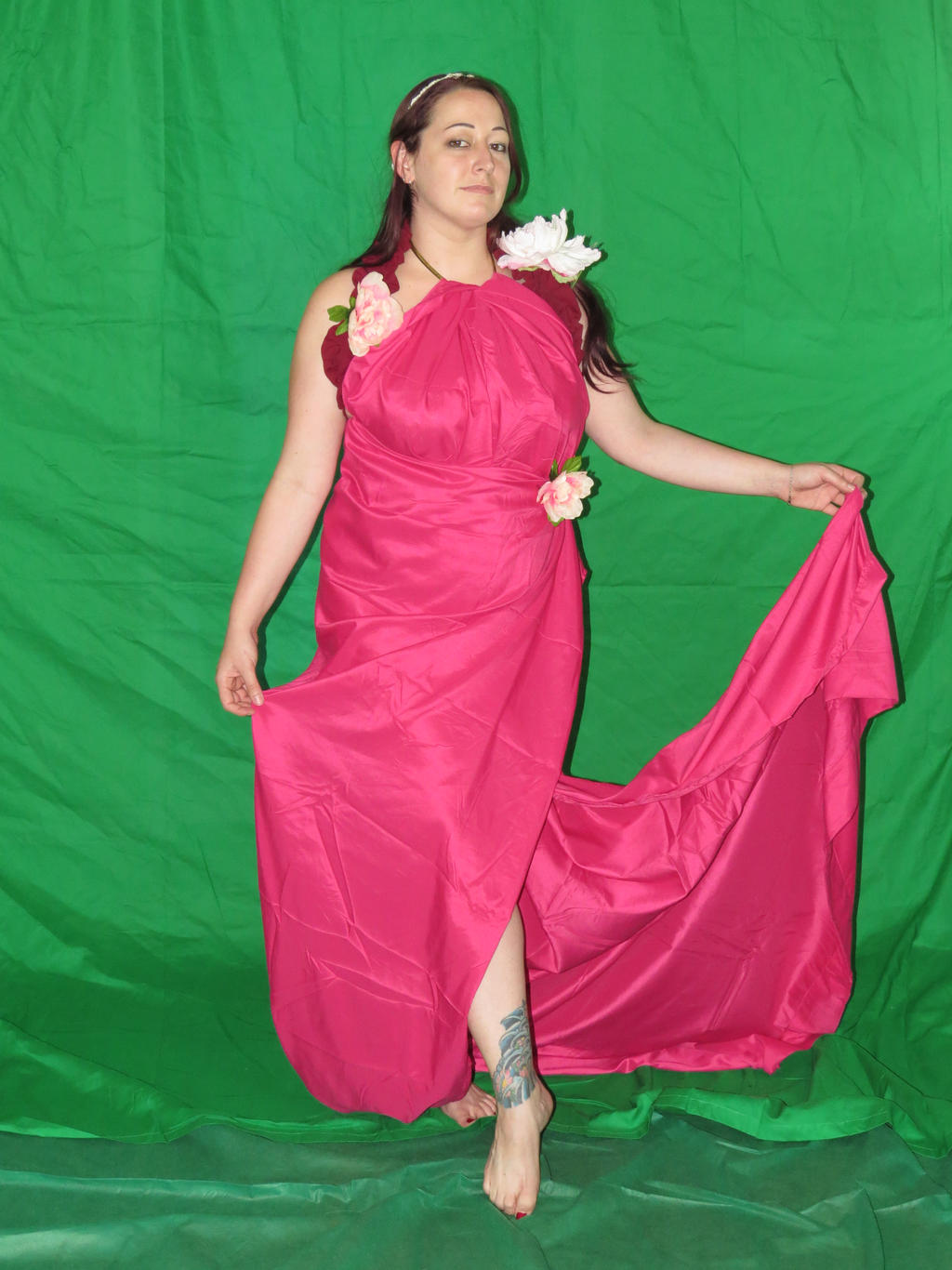 a fulllength portrait of a woman with shoulderlength brown hair wearing a long, flowing, hot pink halterstyle dress adorned with artificial flowers. the dress has a draped design that creates a train effect. she is barefoot, with a visible tattoo on her ankle, and is posing against a bright green backdrop. the overall style suggests a grecian or goddessinspired aesthetic.