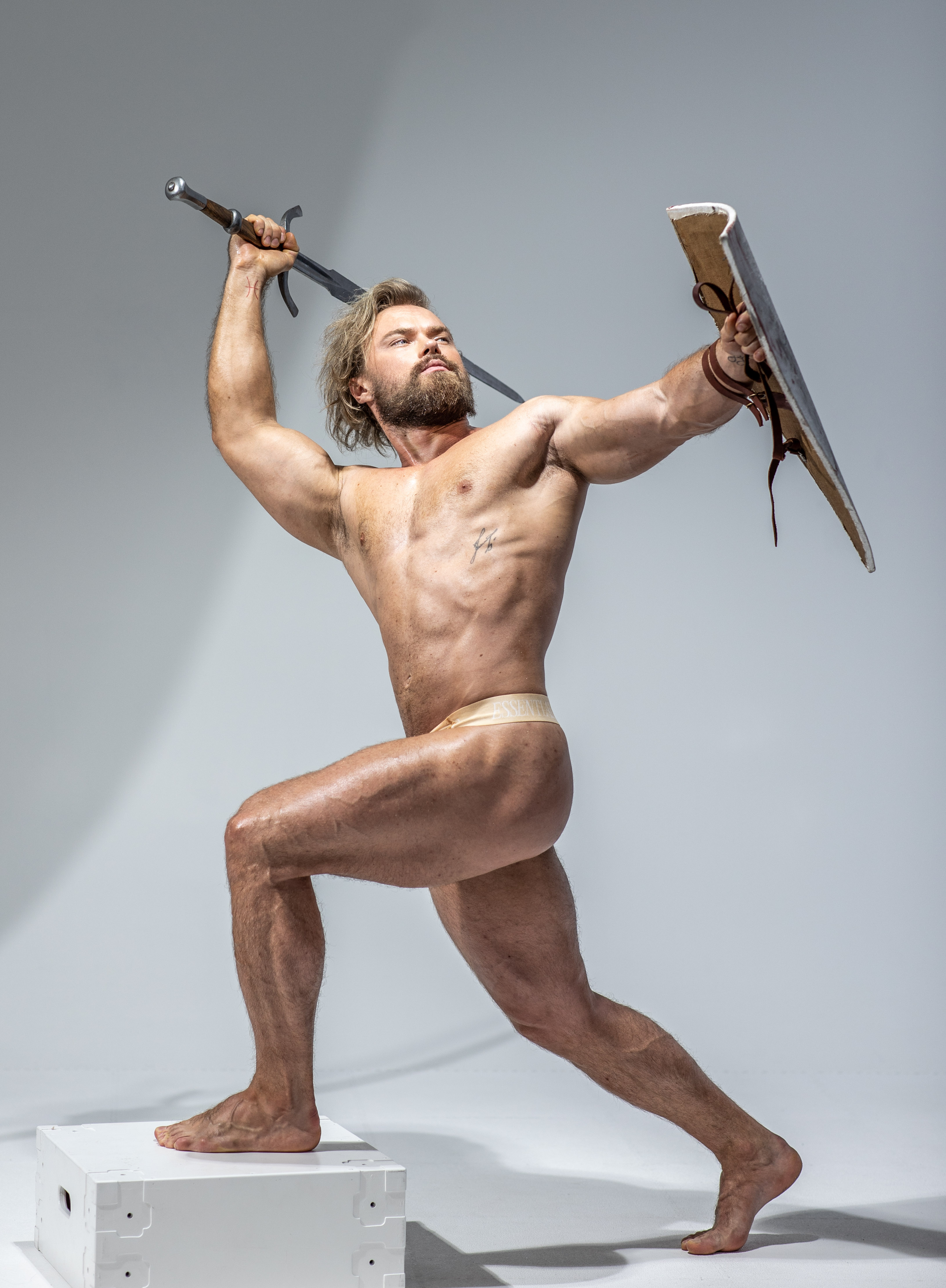 a muscular man with long blond hair poses dynamically against a plain gray background in a studio setting.  he holds a sword aloft in his right hand and a large, rimless shield in his left. he is nude except for a  lightcolored  loincloth. his pose and expression evoke the image of a fierce warrior from history or mythology.  the lighting is bright and highlights his physique.